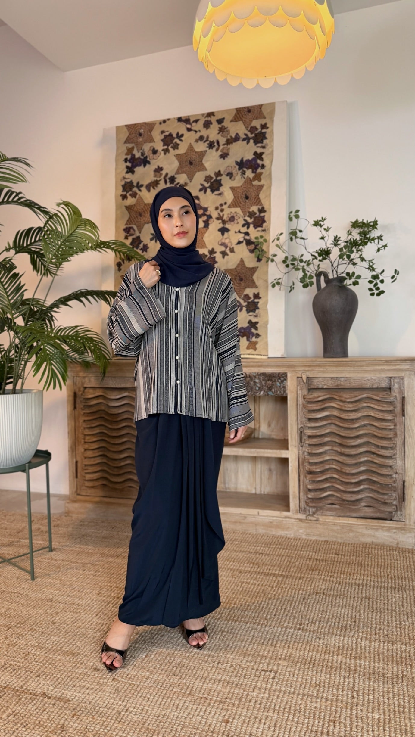 Woman wearing a hijab and long dark skirt in a room with plants and decorative wall art.