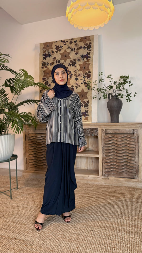 Woman wearing a hijab and long dark skirt in a room with plants and decorative wall art.