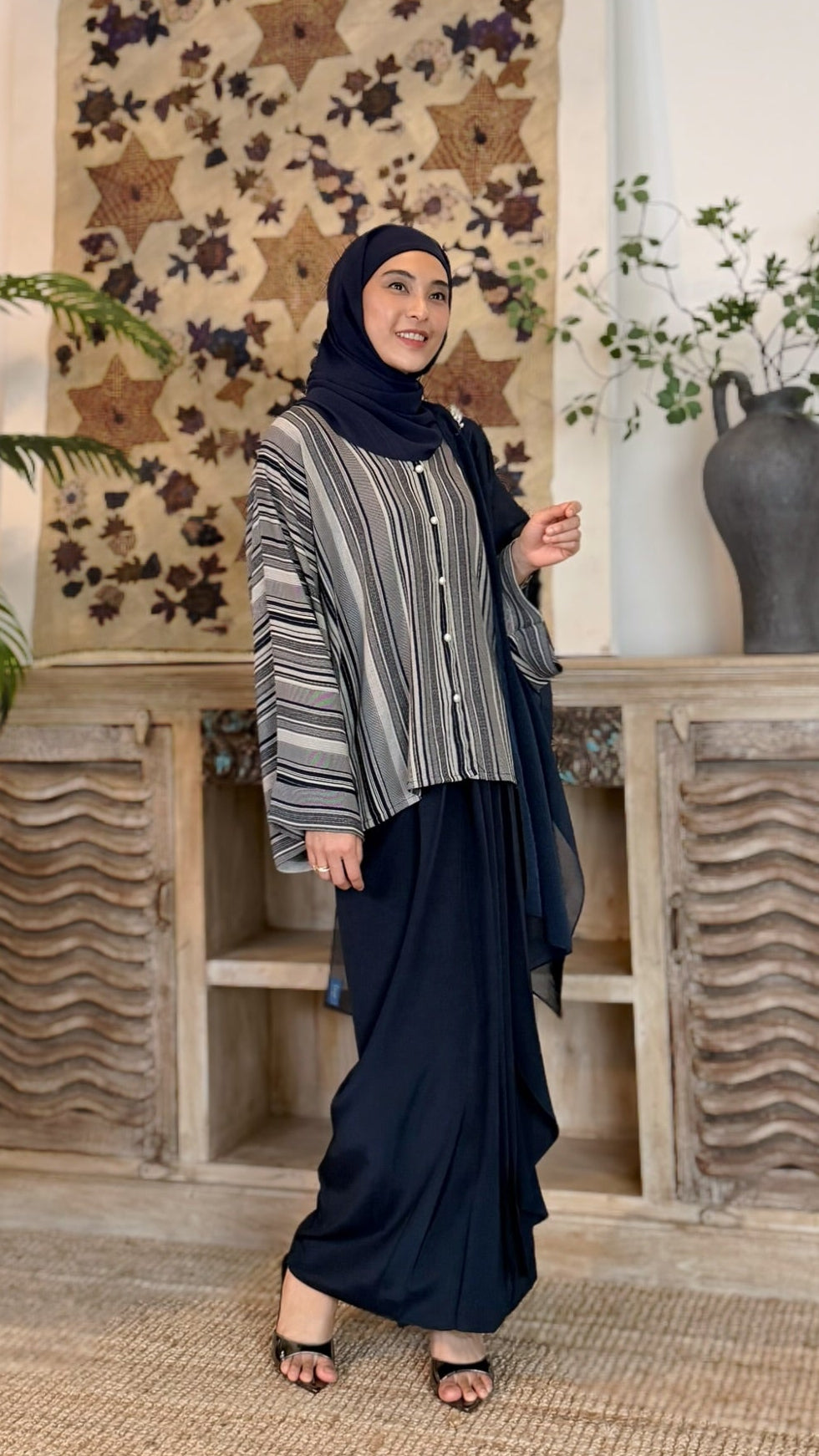 Woman in a hijab standing in a room with decorative wall art and plants.