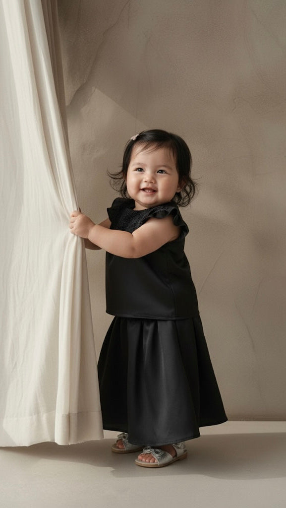 Child in a black dress standing next to a white curtain against a beige wall.