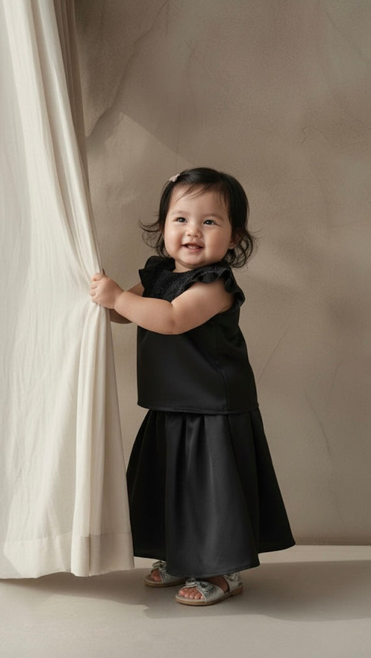 Child in a black dress standing next to a white curtain against a beige wall.