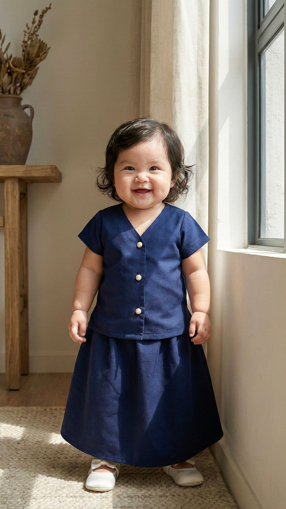 Child wearing a navy outfit standing in a room with a window and wooden table.
