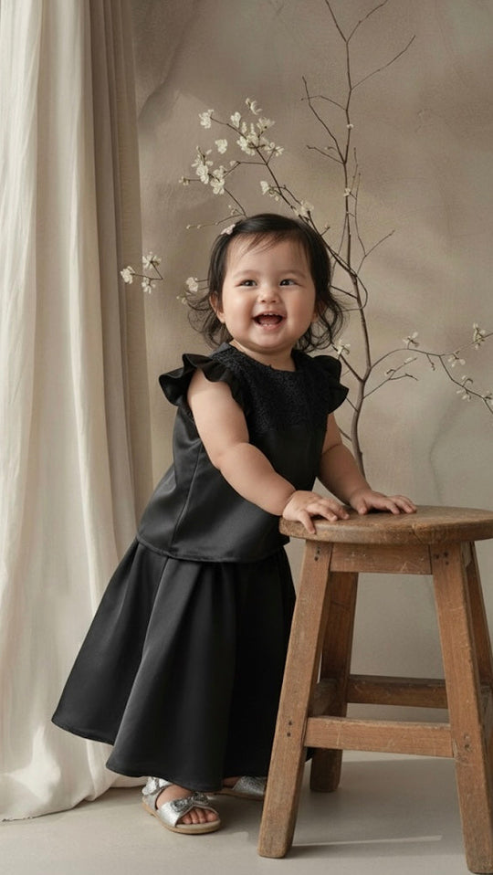Child in a black dress standing on a wooden stool with white curtains and branches in the background